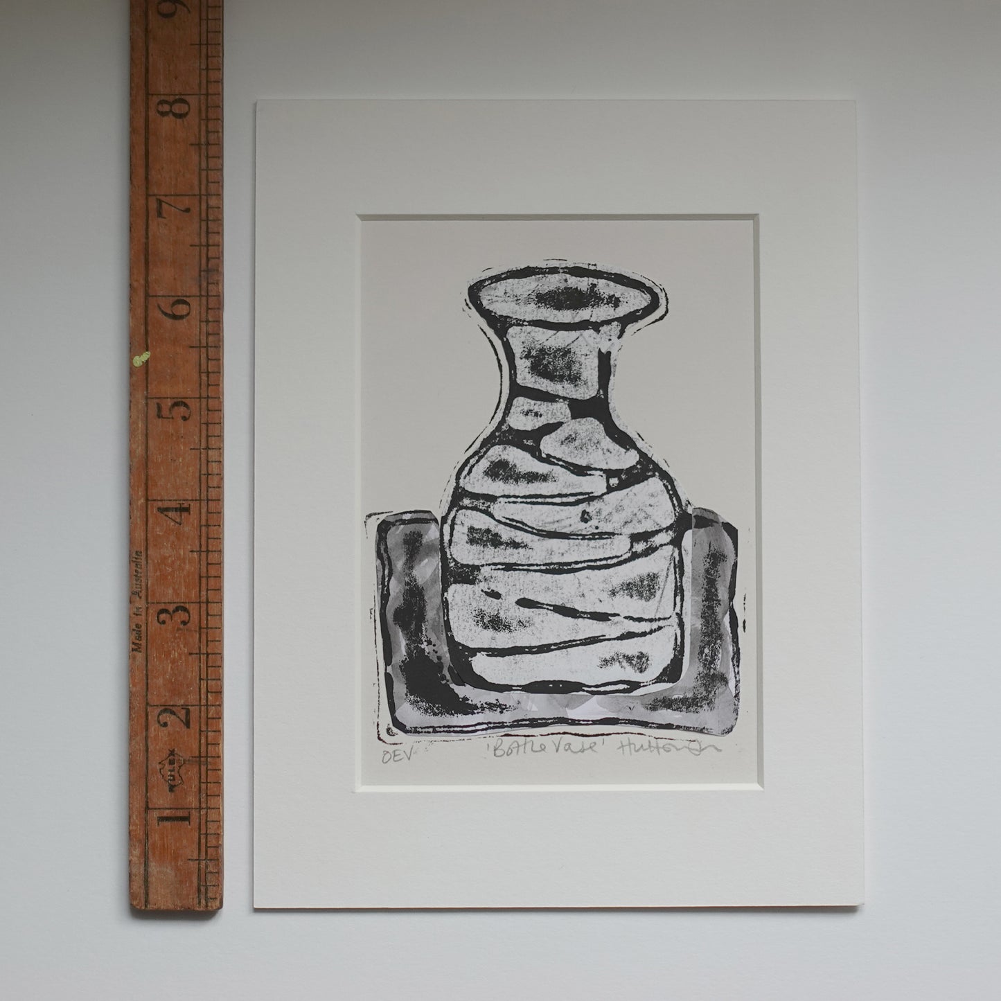 Relief print of bottle vase printed with black ink over chine-collé shown in mount with wooden ruler for scale